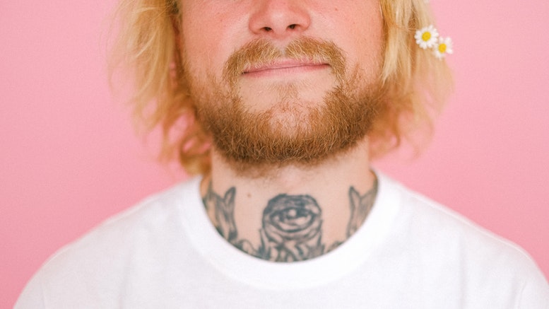 Bottom half of a persons face, showing facial hair, a tattoo on the neck, wearing a white shirt.